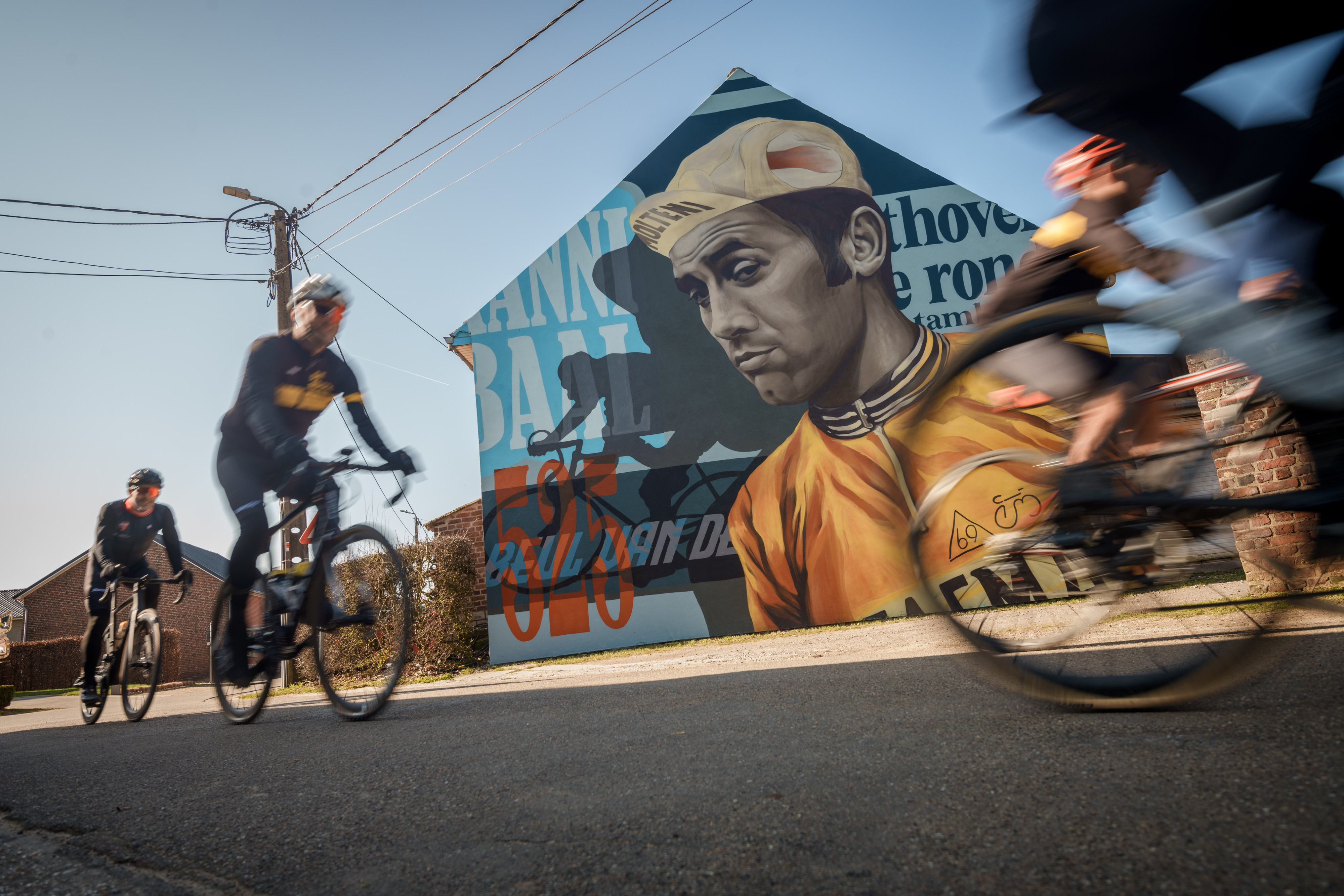 Afbeelding van de muurschildering van Eddy Merckx met voorbijrijdende fietsers op de voorgrond.